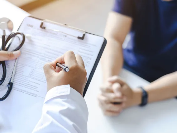 close up of doctor's hand writing on a prescription notepad with patient hands clasped in the background