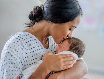 mother in hospital gown cradling and kissing forehead of swaddled infant