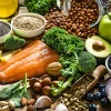 Healthy foods on a table: salmon, kale, spinach, avocado, almonds, blueberries, onion, chia seeds, cucumber, tomato, apple, pistachios, garlic, carafe of olive oil, lemon, broccoli